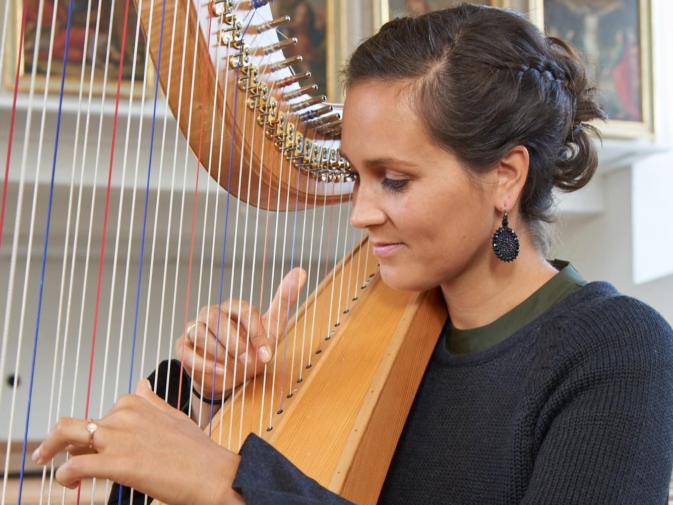 Eine Frau spielt Harfe in einem hellen Raum. Sie trägt elegante Ohrringe und konzentriert sich auf die Musik.