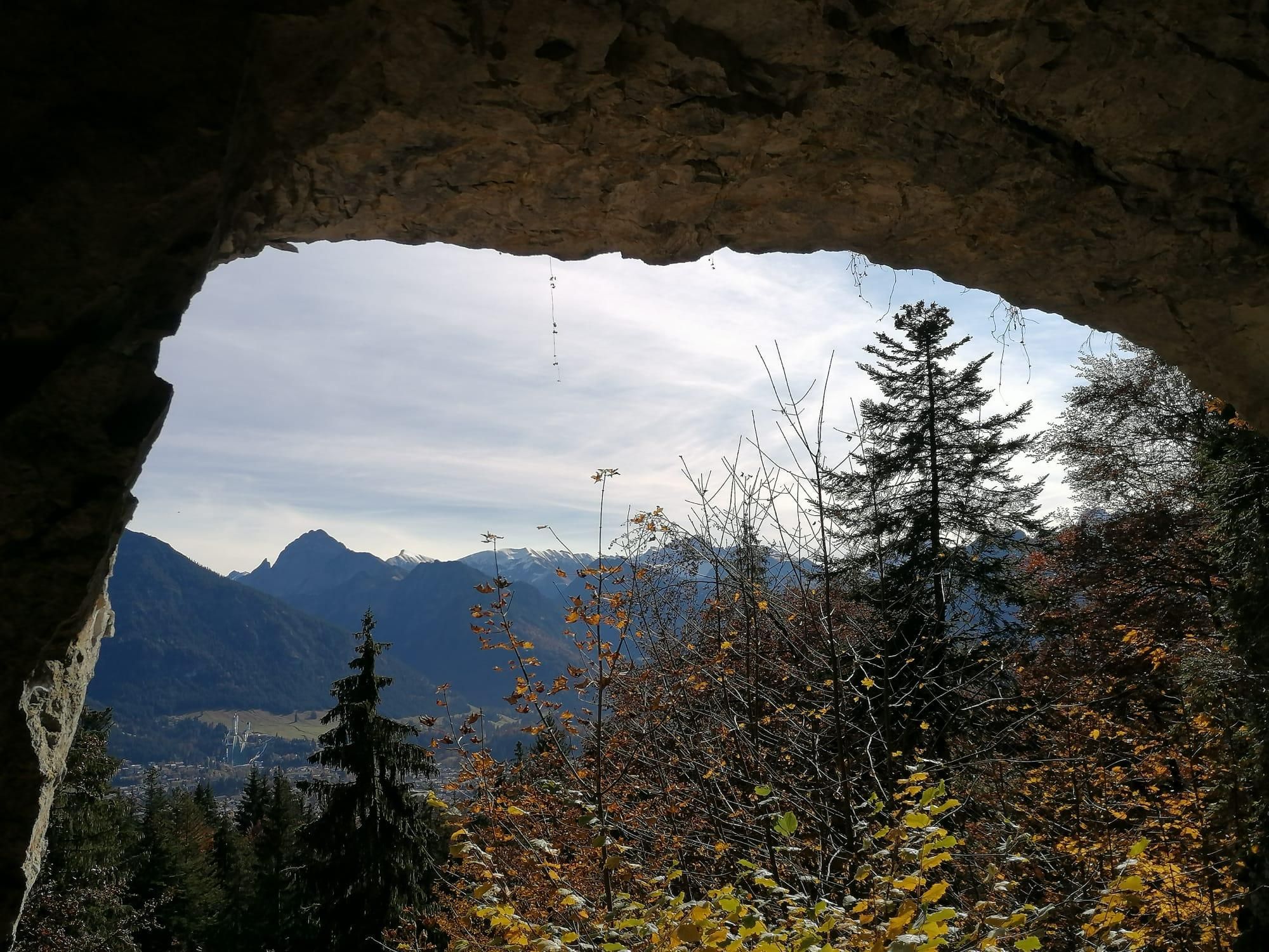 Geräumiges Felsentor mit Blick auf ein Bergpanorama. Der Himmel ist hellblau mit einigen Wolken. Im Vordergrund herbstlich gefärbte Bäume und Sträucher.