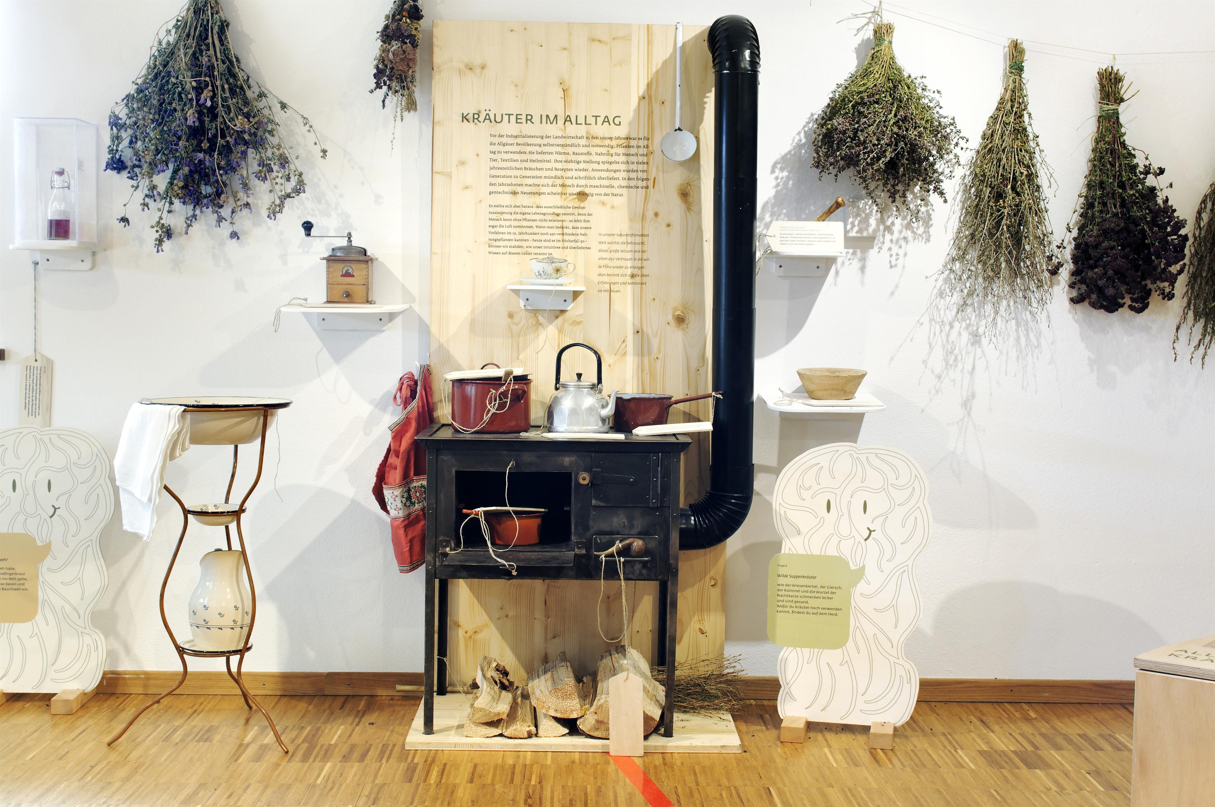 Ausstellungsstück: ein alter Holzofen mit Ofenrohr, Kräuterbündel an der Wand, links eine Waschschüssel mit Krug.