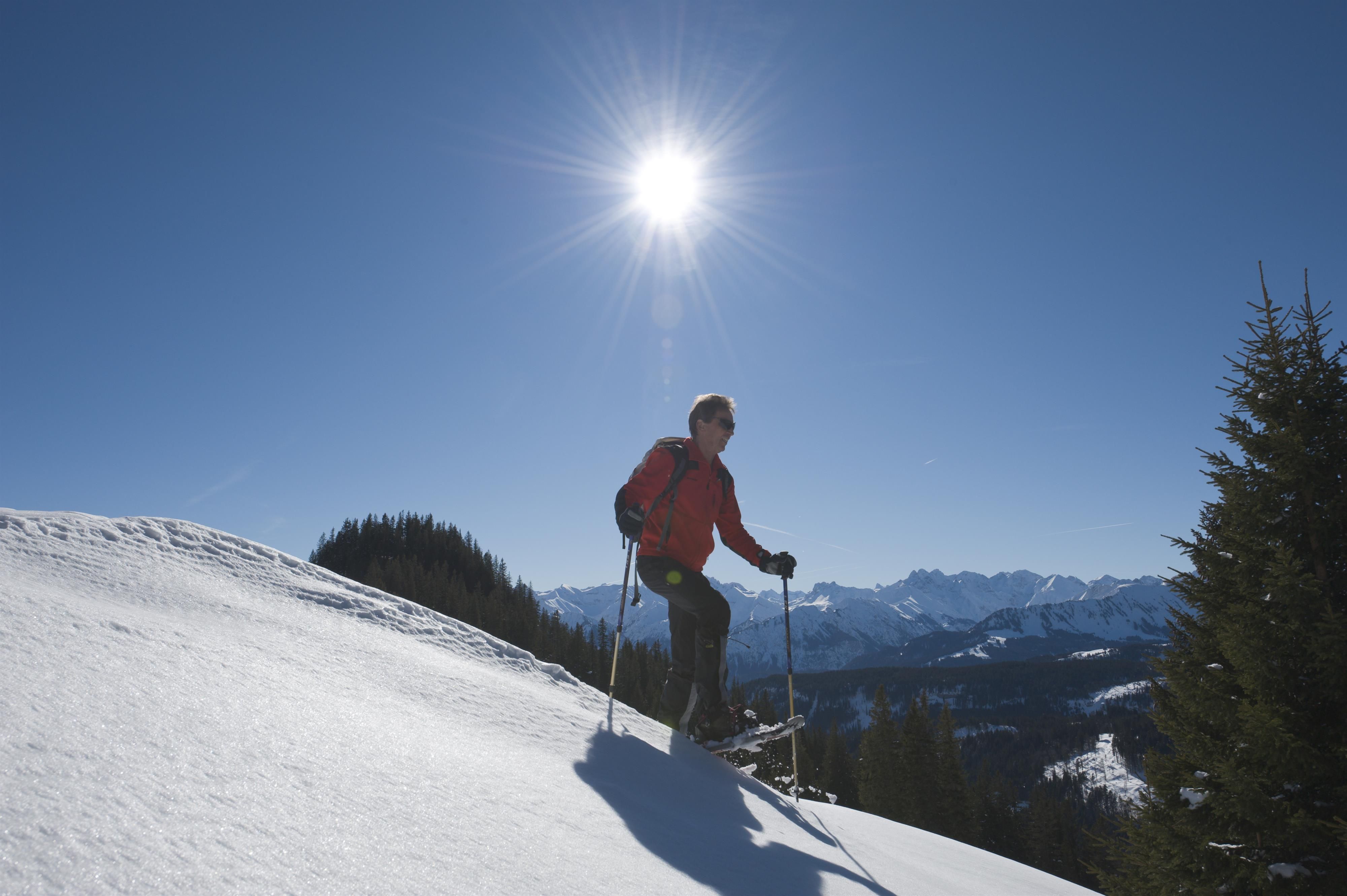 Ein Mann mit Skibekleidung läuft an einem sonnigen Tag mit Schneeschuhen über einen verschneiten Hügel.