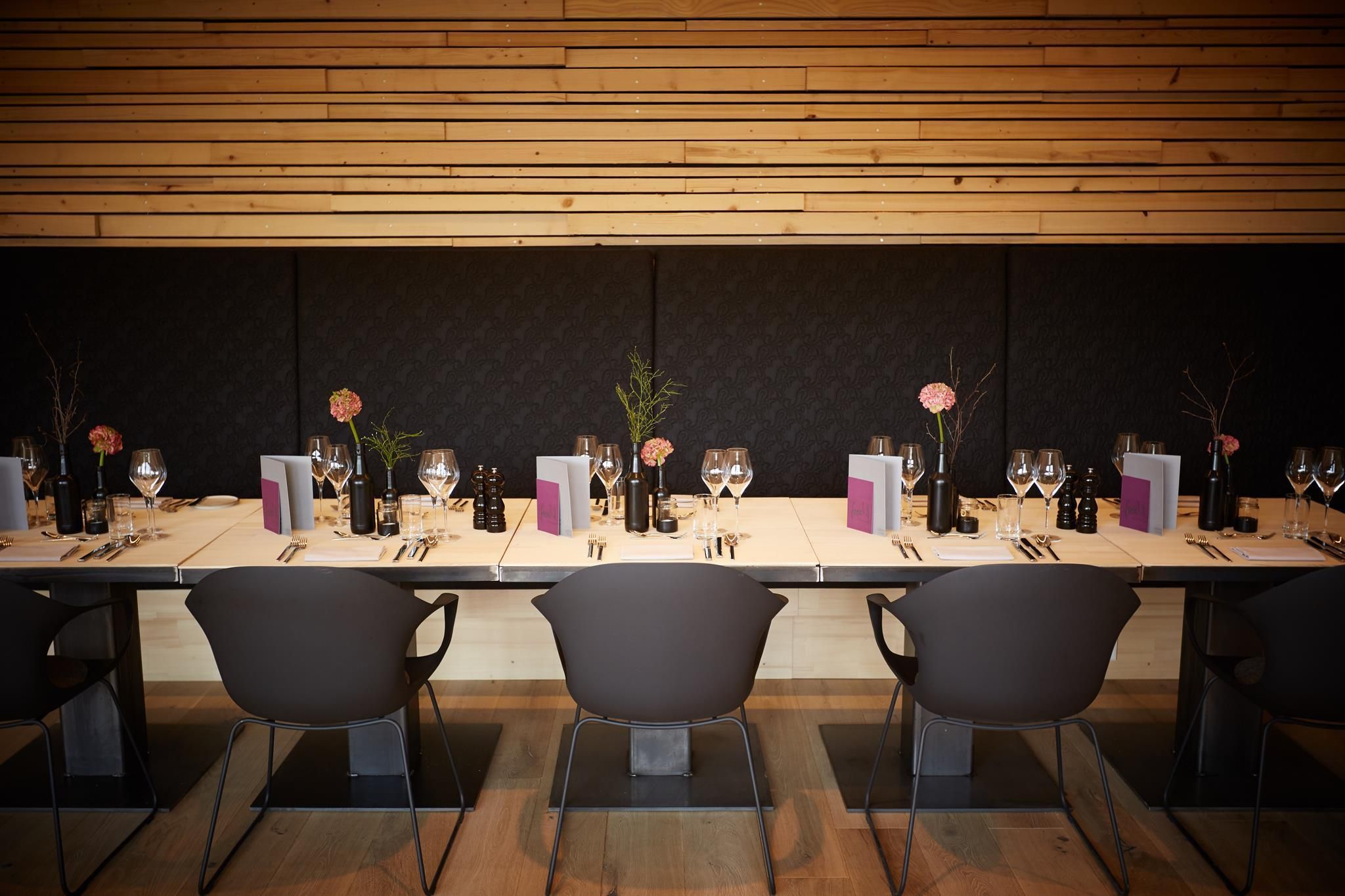 Restaurant Freistil Ofterschwang: Langer Holztisch, graue Stühle, moderne Dekoration mit rosa Blüten. Holz und Stoff an der Wand. Helle Böden.