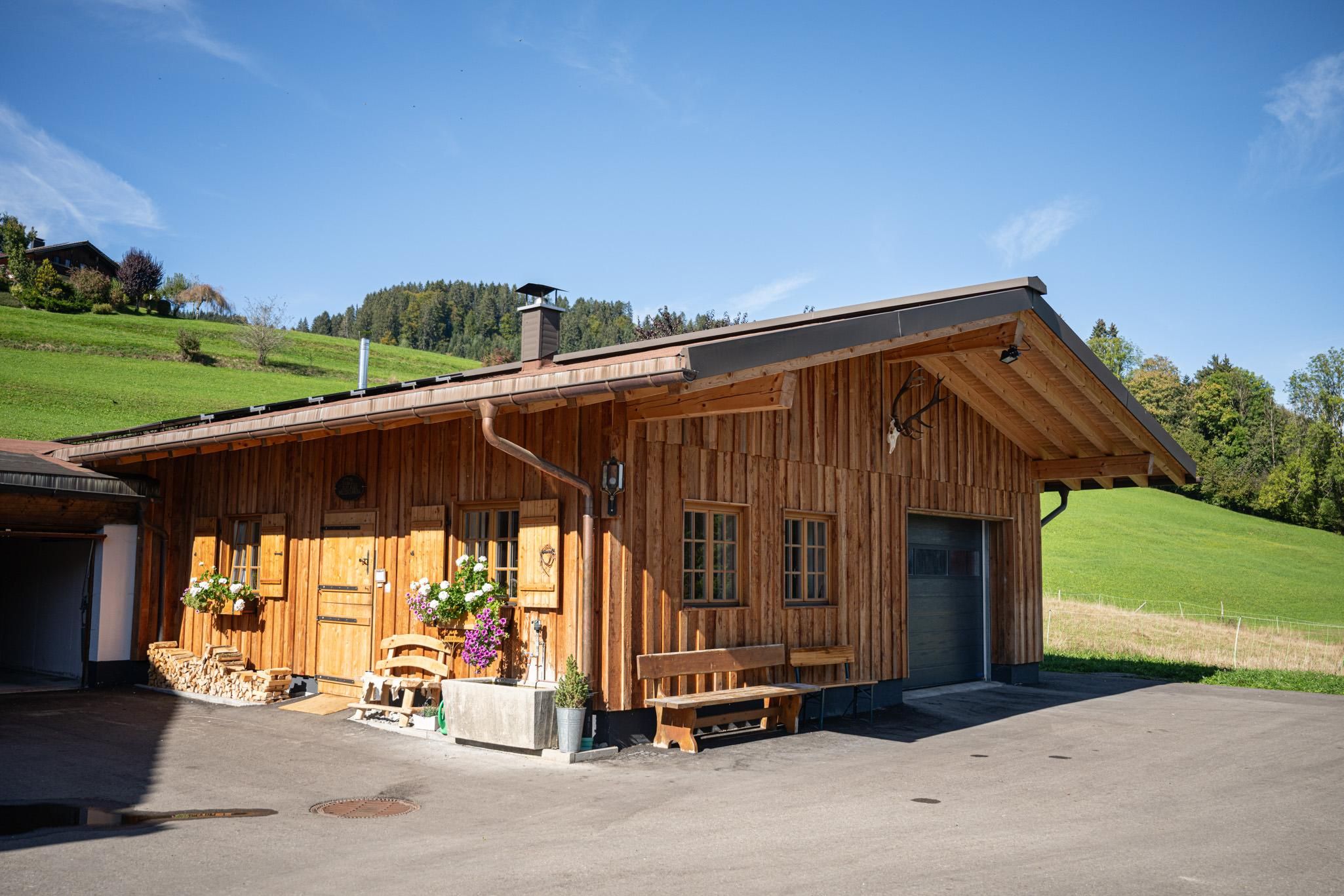 Holzhaus mit Satteldach in ländlicher Umgebung, grüne Wiesen und blauer Himmel.