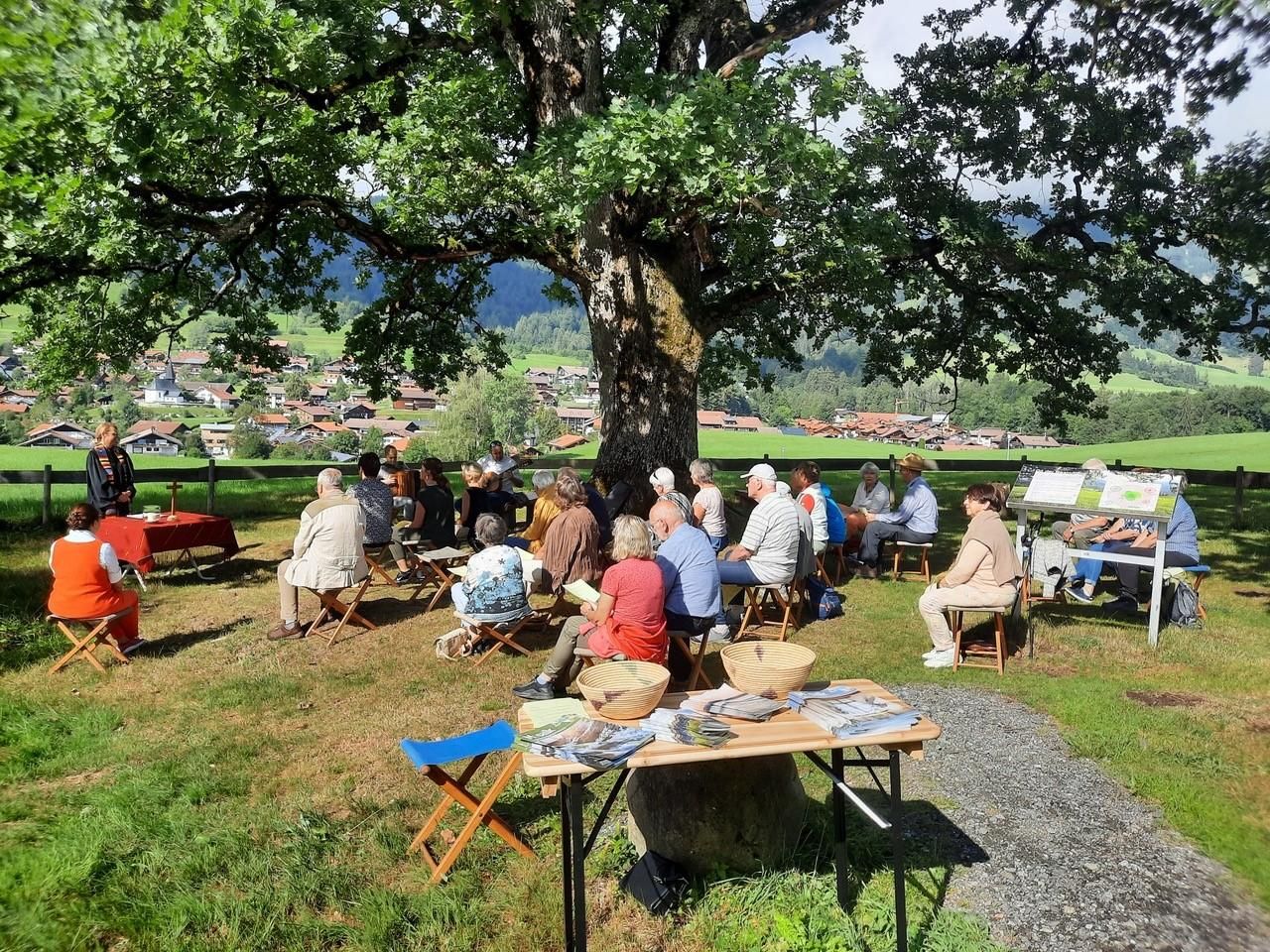 Eine Gruppe von Menschen sitzt bei einem Gottesdienst im Freien unter einem großen Baum. Im Hintergrund sind Wiesen und Berge sowie einige Häuser zu sehen.