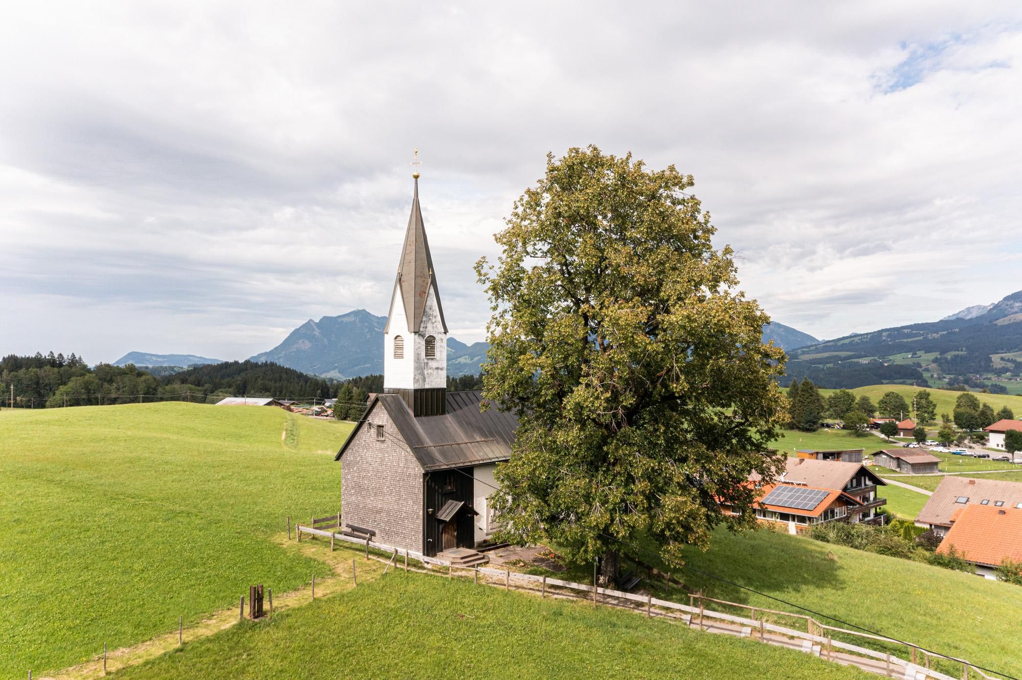 Kleine Kapelle. Graues Gebäude mit spitzem, hellem Turm. Steht auf einer grünen Wiese neben einem großen Baum. Berge im Hintergrund.