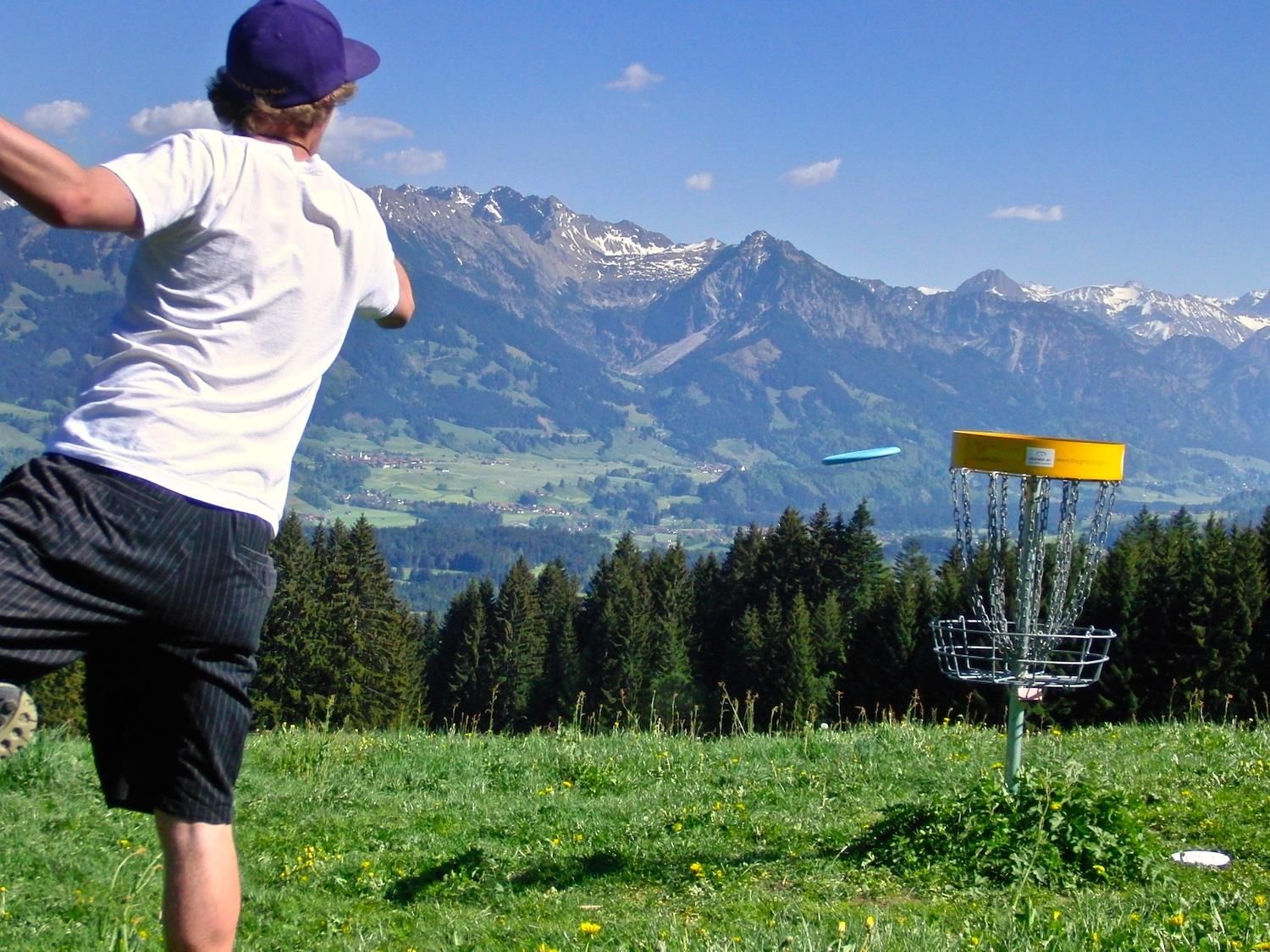 Mann wirft blaue Frisbeescheibe auf gelben Disc Golf Korb.