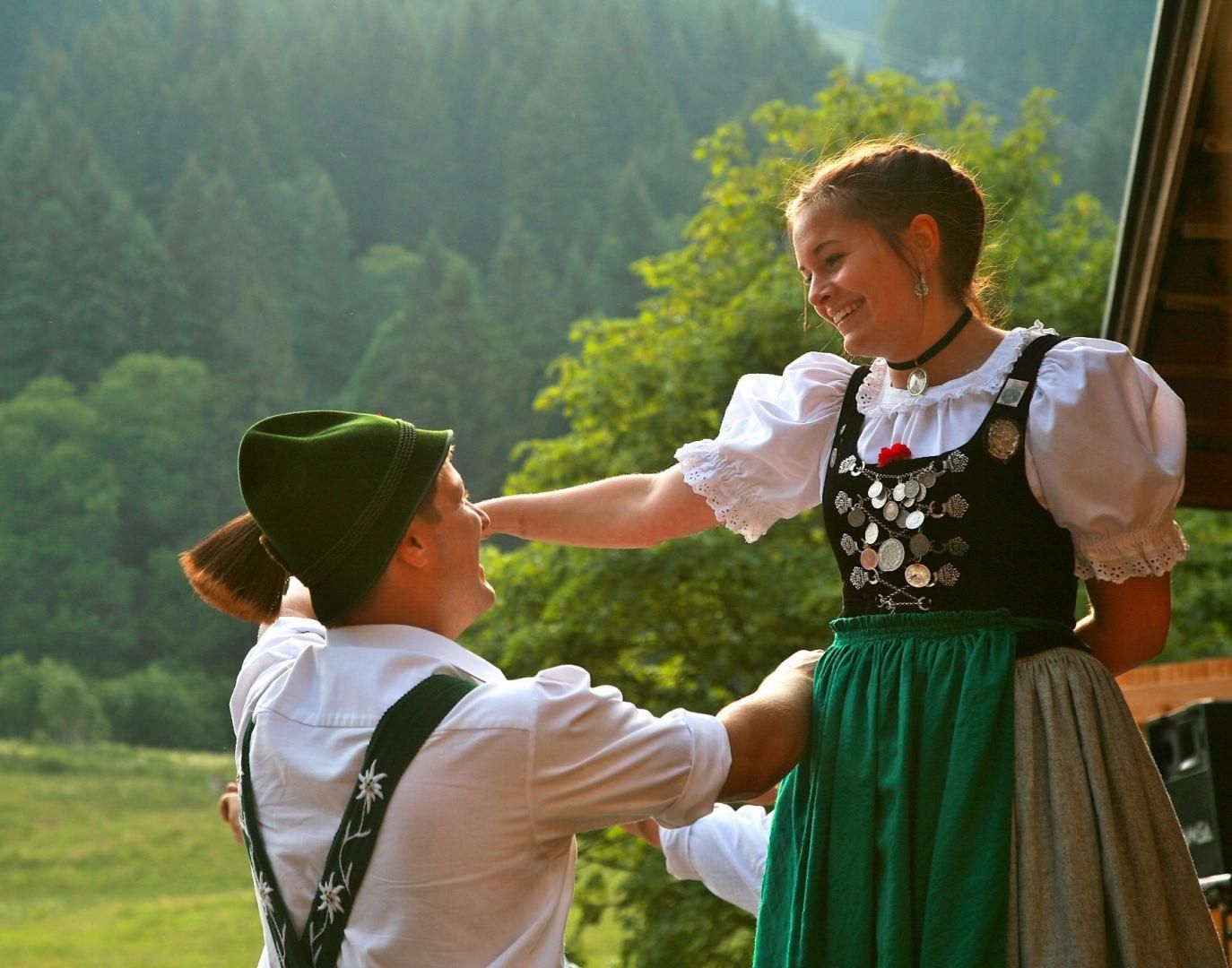 Junges Trachtenpaar beim Volkstanz. Sie lächelt, im Dirndl. Er trägt Hemd, Hosenträger, grünen Hut.