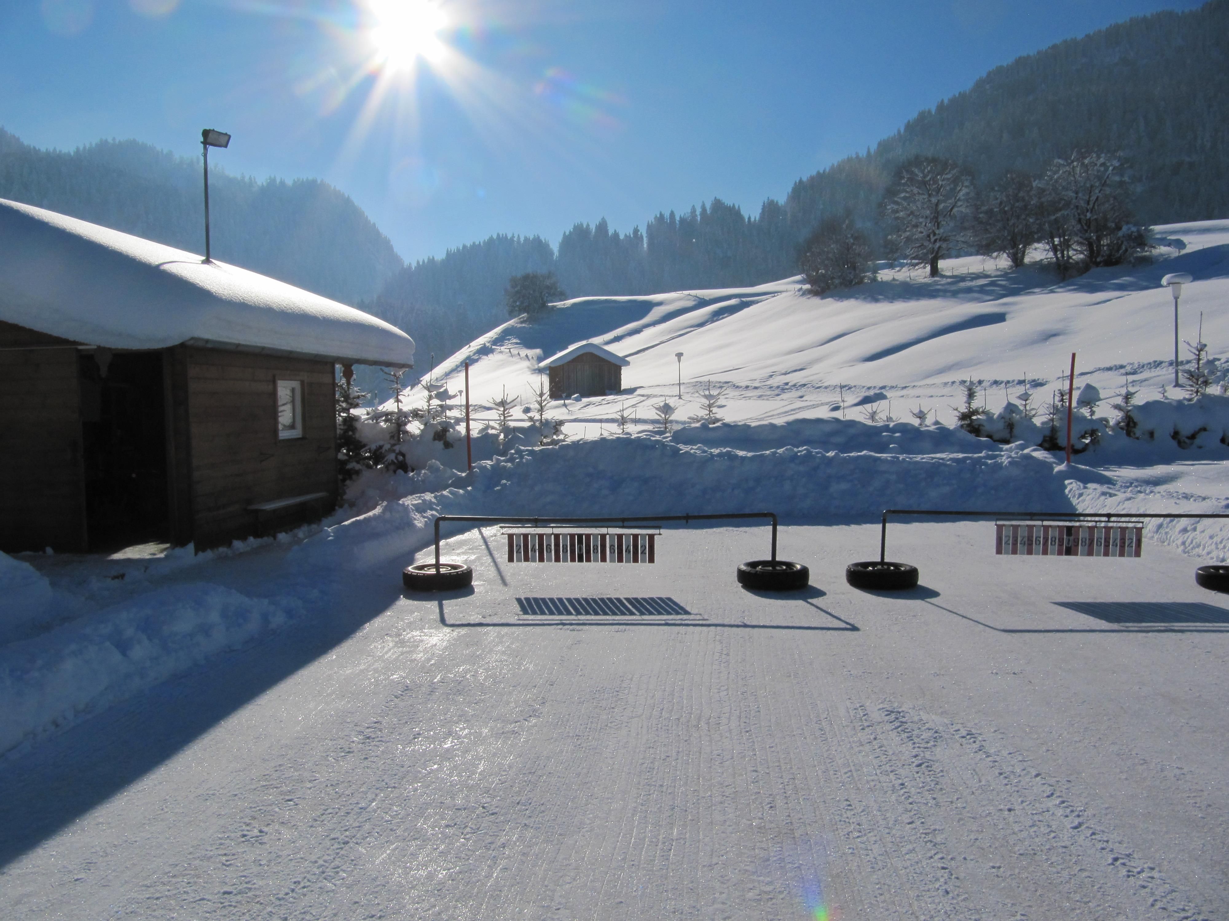 Sonniger Wintertag auf einem Eisstockplatz mit schneebedeckten Bergen und Hütten.