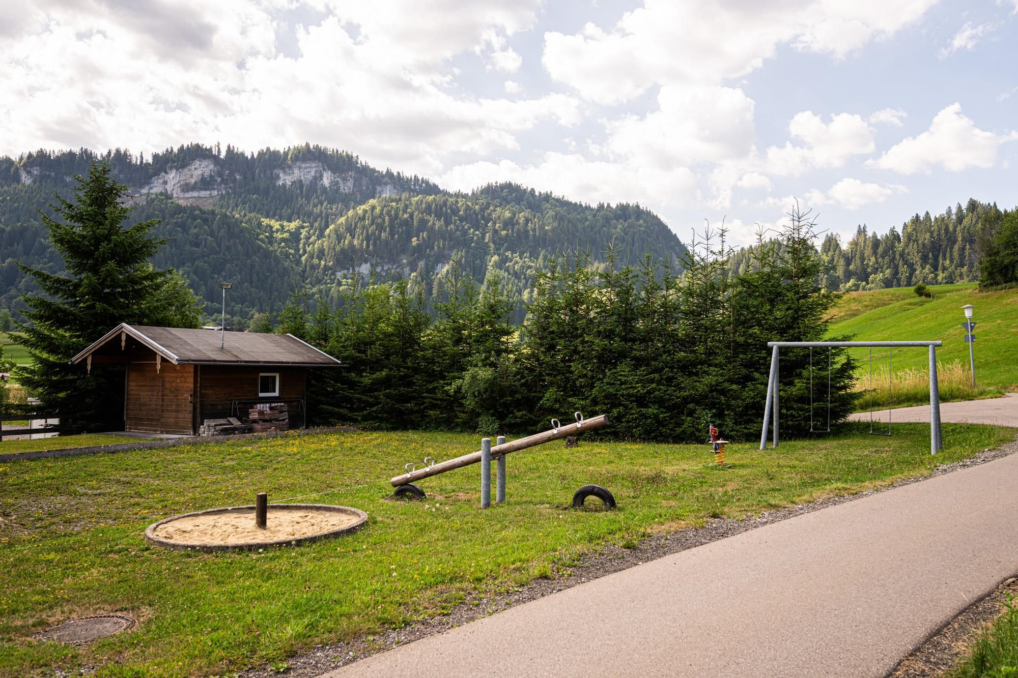 Spielplatz Rieder Wiesen. Holzhütte, Sandkasten, Wippe und Schaukel auf grüner Wiese. Befestigter Weg führt vorbei.