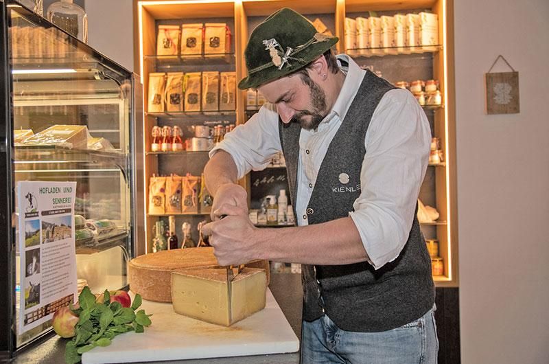 Mann mit Bart und Trachtenhut zerteilt einen großen Käseblock mit einem Käsemesser. Hinter ihm Regale mit verschiedenen Produkten und eine Kühltheke.