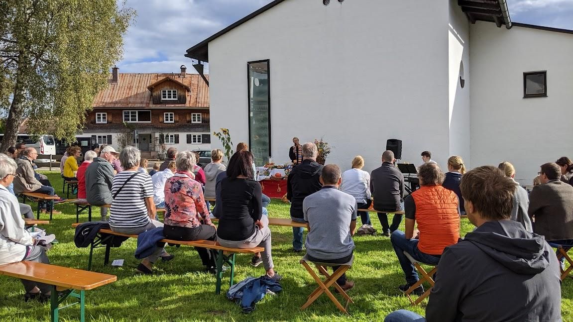 Eine Gruppe von Menschen hört der Pfarrerin im Freien zu. Im Hintergrund sind mehrere Gebäude und Bäume zu sehen.