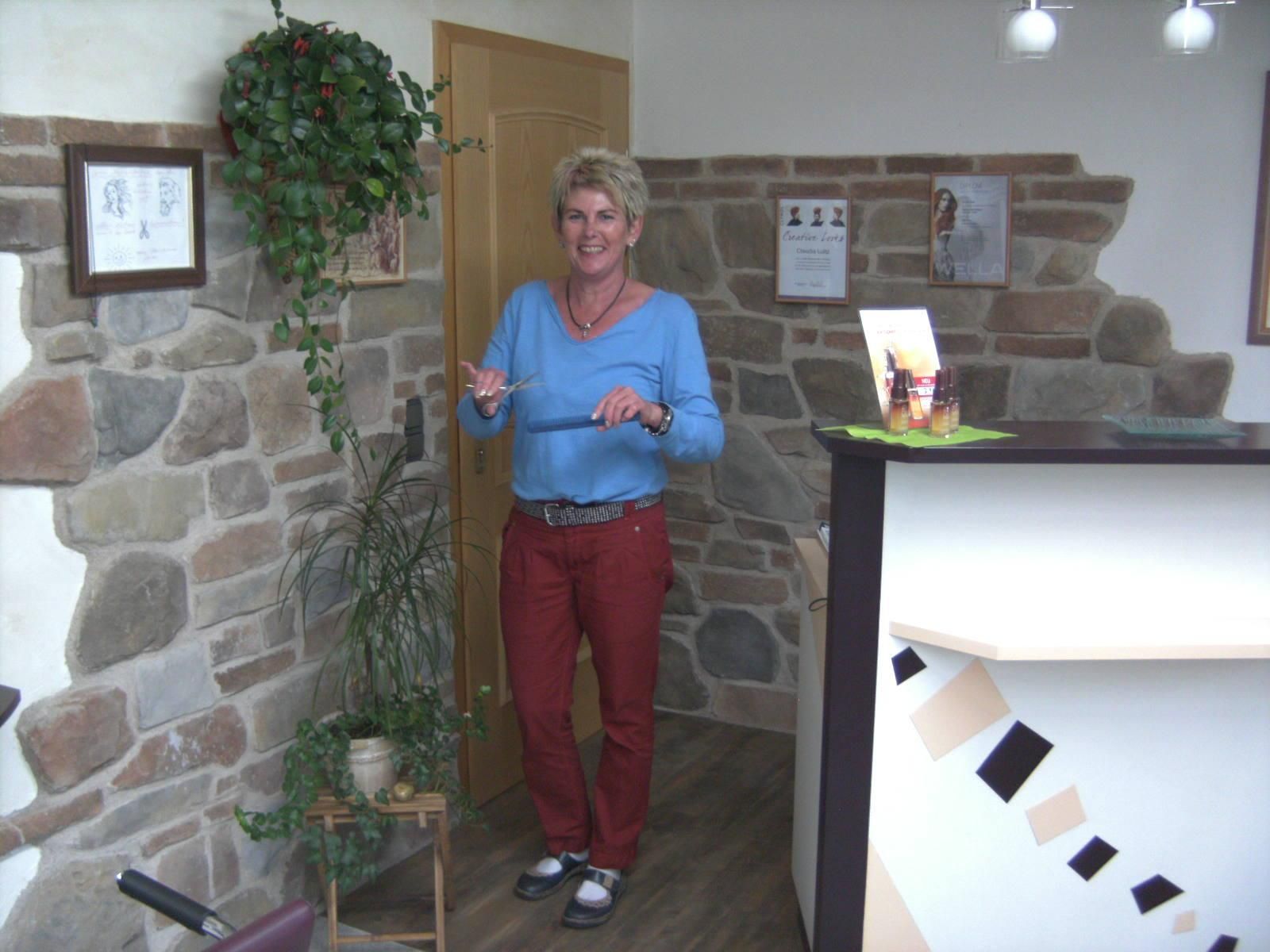 Eine freundliche Frau mit kurzen blonden Haaren steht im Eingangsbereich des Friseursalons Haarmonie.