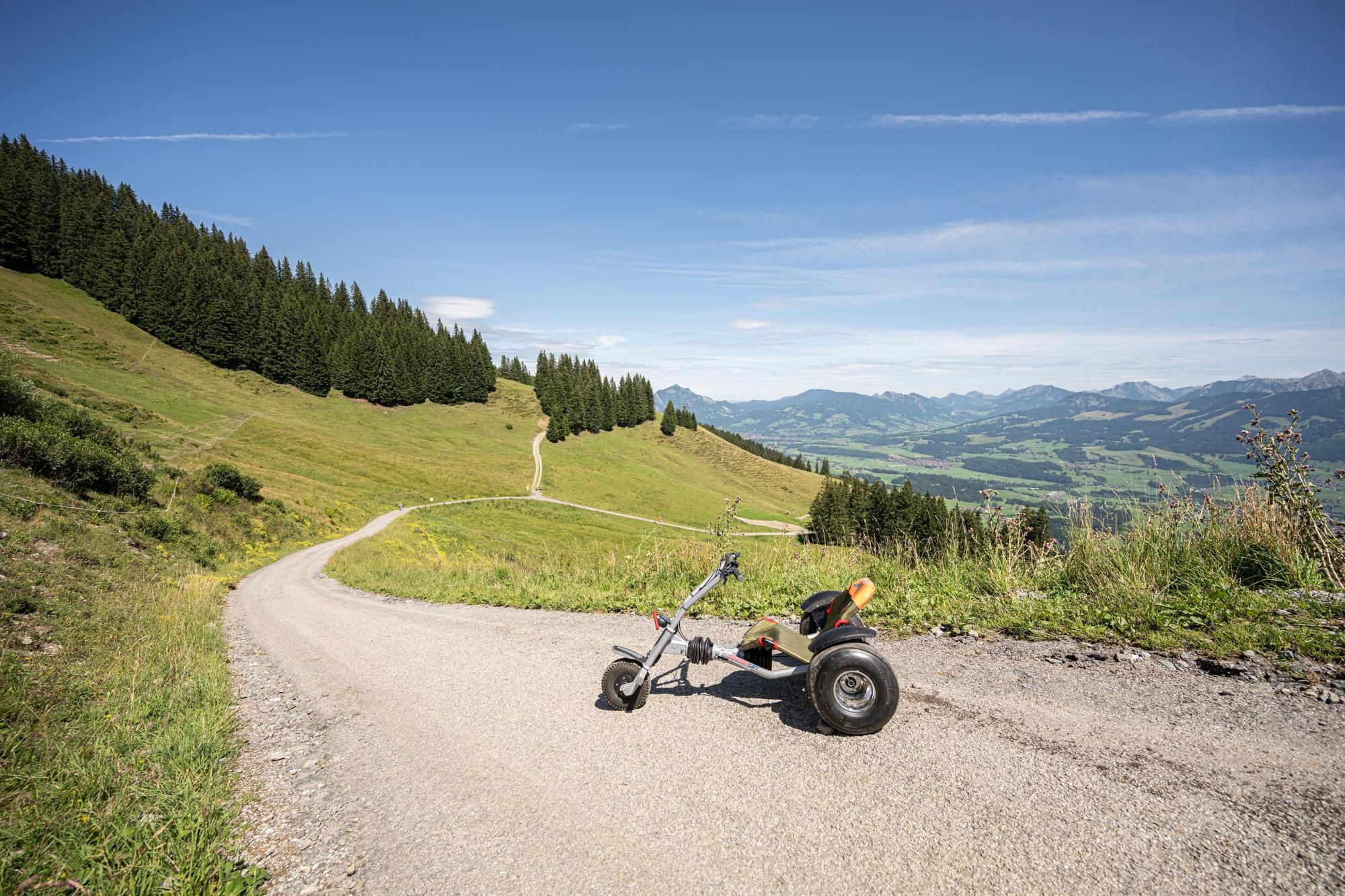 Mountaincart in Berglandschaft: Dreirad mit Sitz auf Schotterweg, grüne Hänge mit Bäumen, Bergpanorama unter blauem Himmel.