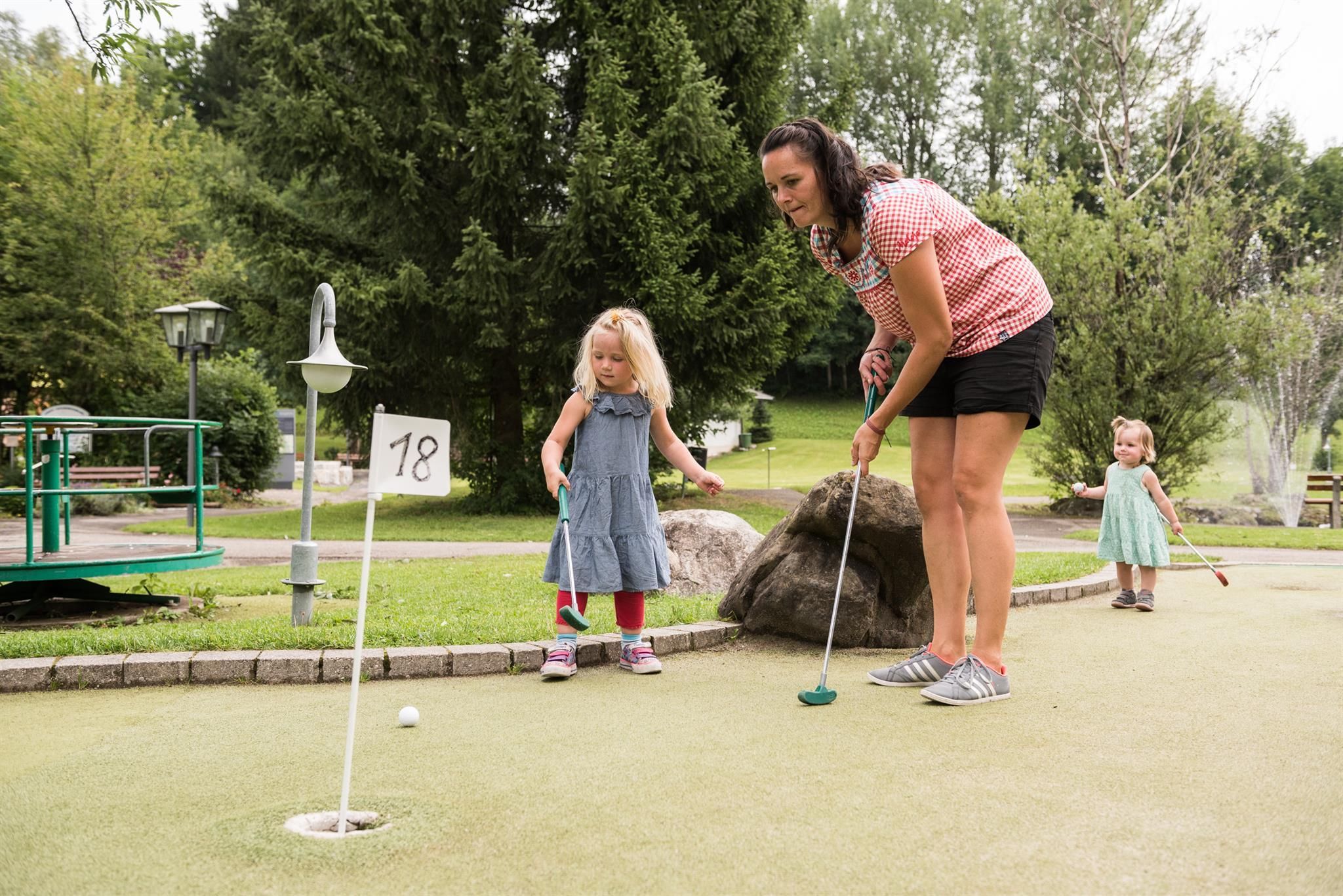 Grüne Minigolfanlage in Obermaiselstein. Mehrere Bahnen mit unterschiedlichen Hindernissen. Bunte Markierungen an den Bahnen. Umgeben von grüner Wiese und Bäumen.