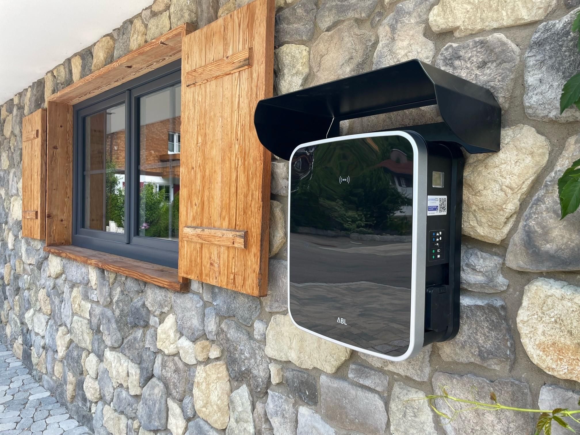 Eine moderne, schwarze E-Auto-Ladestation mit weißer Umrandung ist an einer steinernen Hauswand neben einem Fenster mit Holzläden montiert.