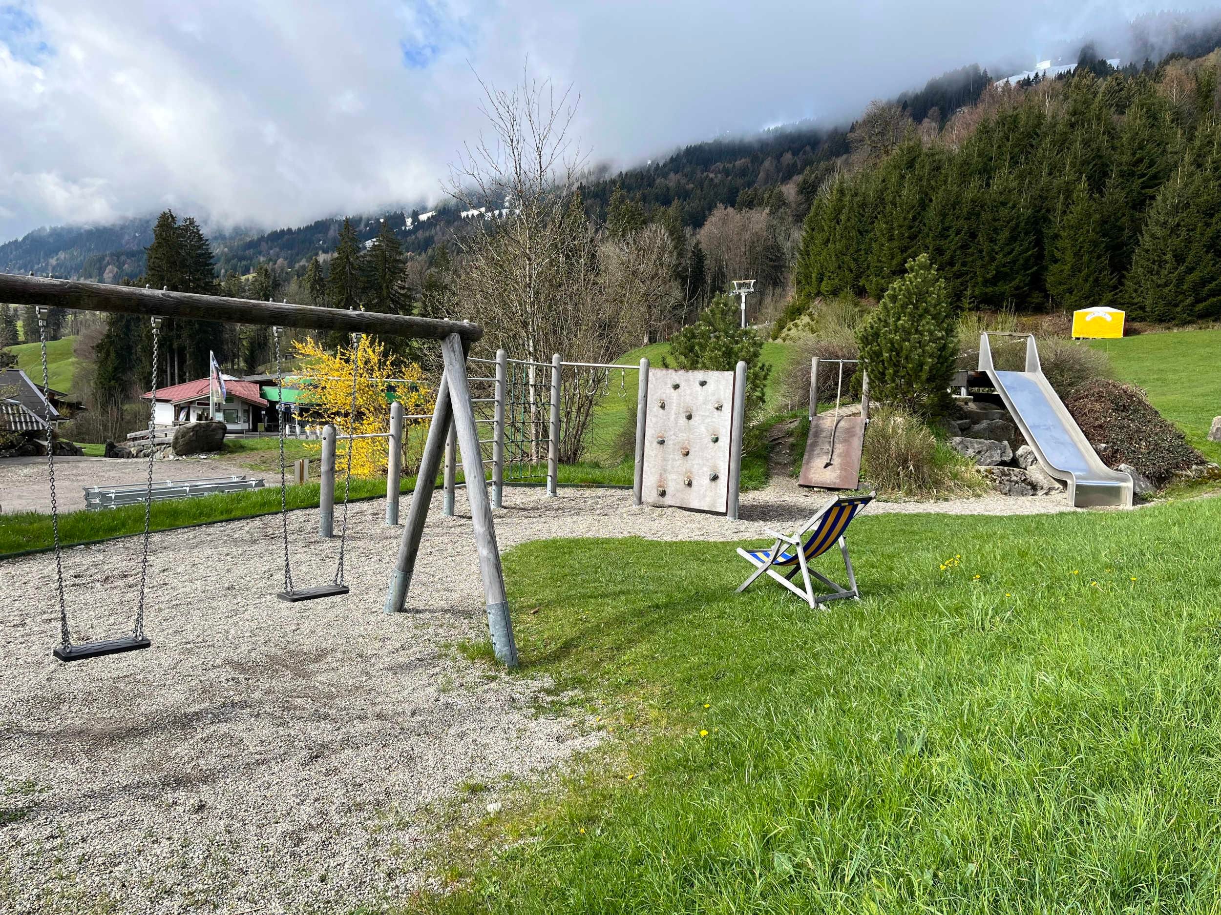 Spielplatz mit Schaukeln, Kletterwand, Rutsche und einer Liege auf einer grünen Wiese. Kiesboden unter den Spielgeräten. Bewaldeter Hang und bewölkter Himmel im Hintergrund.