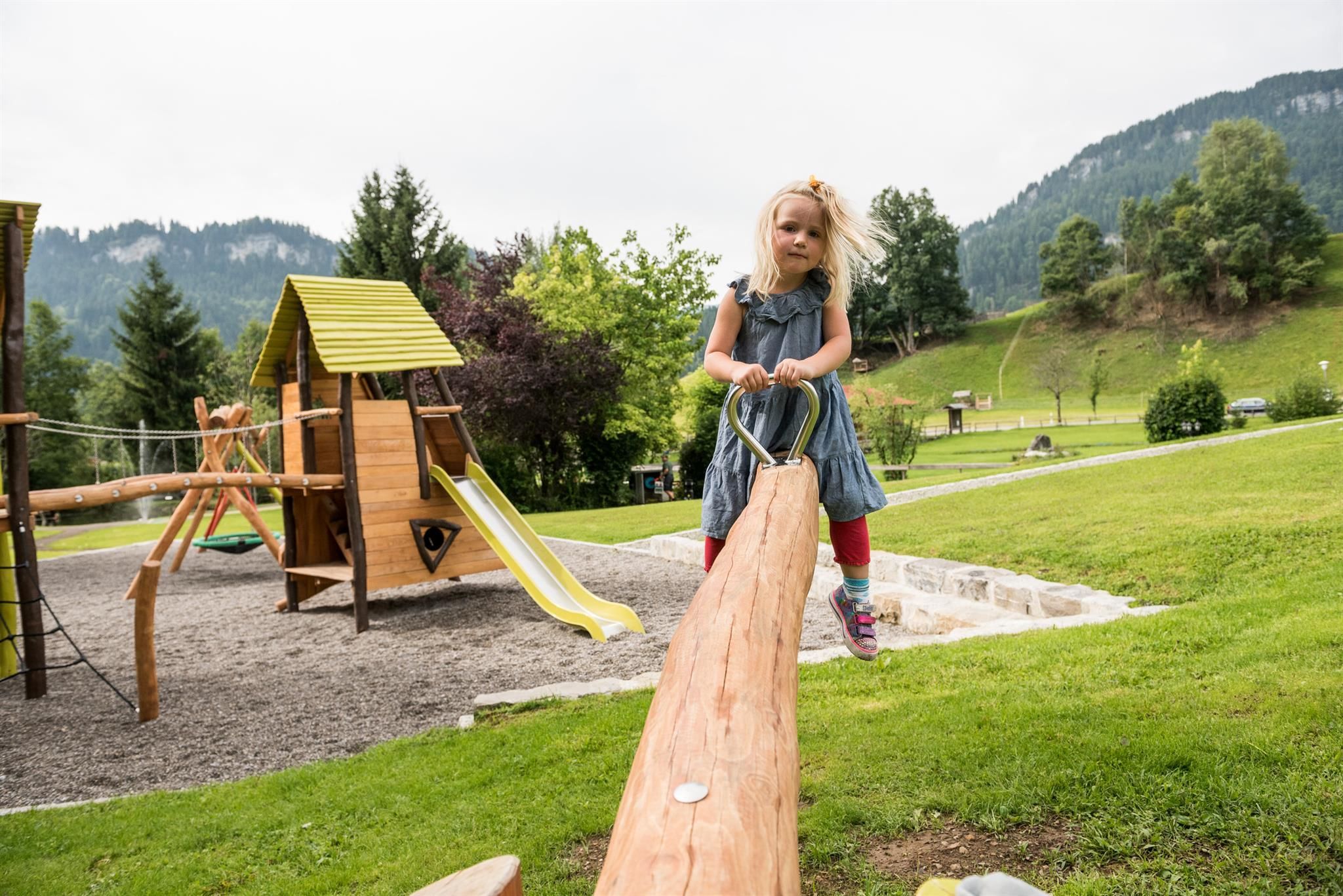 Mädchen mit blonden Haaren auf einer hölzernen Wippe auf einem Spielplatz, im Hintergrund Spielgeräte, grüne Wiese und bewaldete Berge.