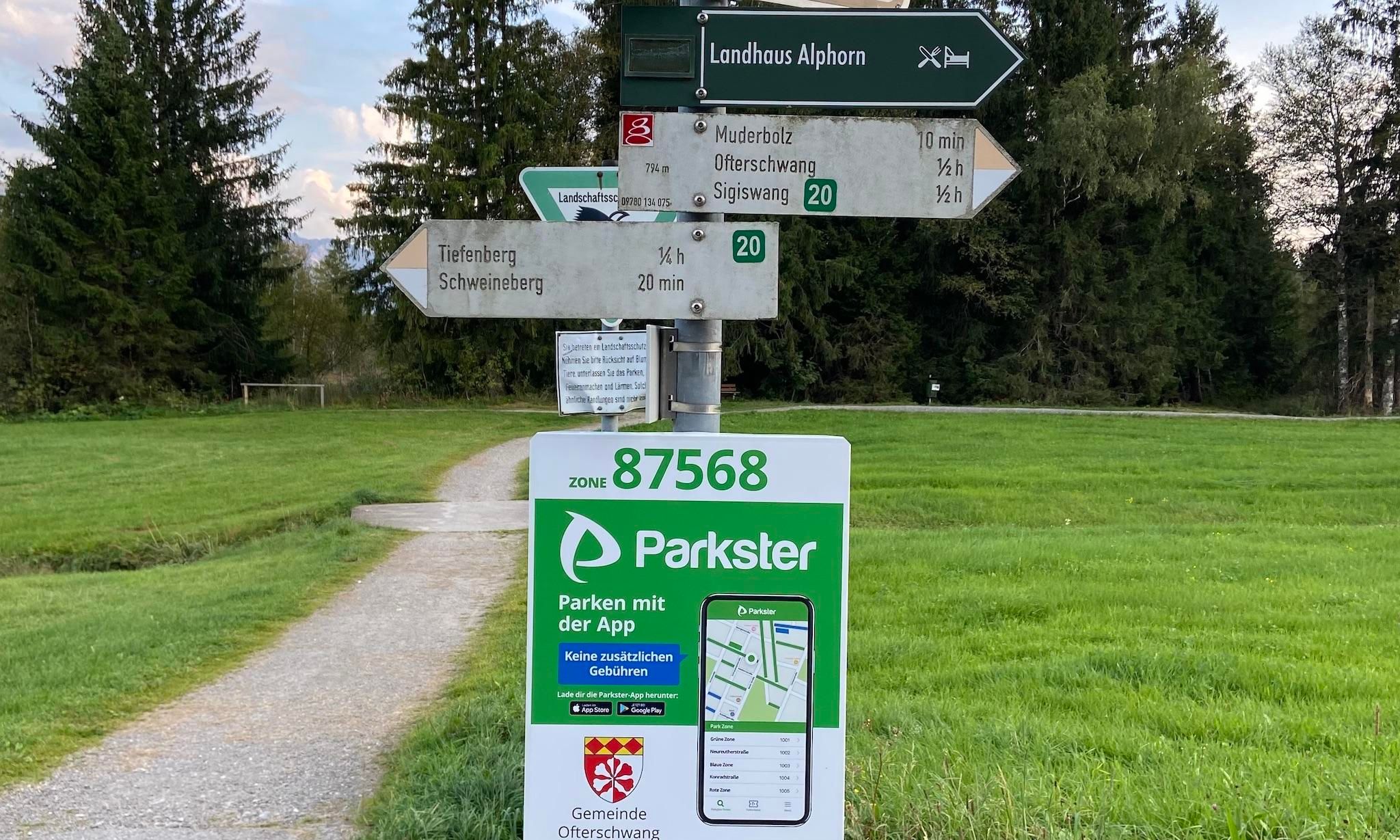 Wegweiser mit. Darunter ein Parkster Schild mit QR-Code und App-Hinweis "Parken mit der App". Schotterweg führt in grüne Landschaft mit Bäumen.