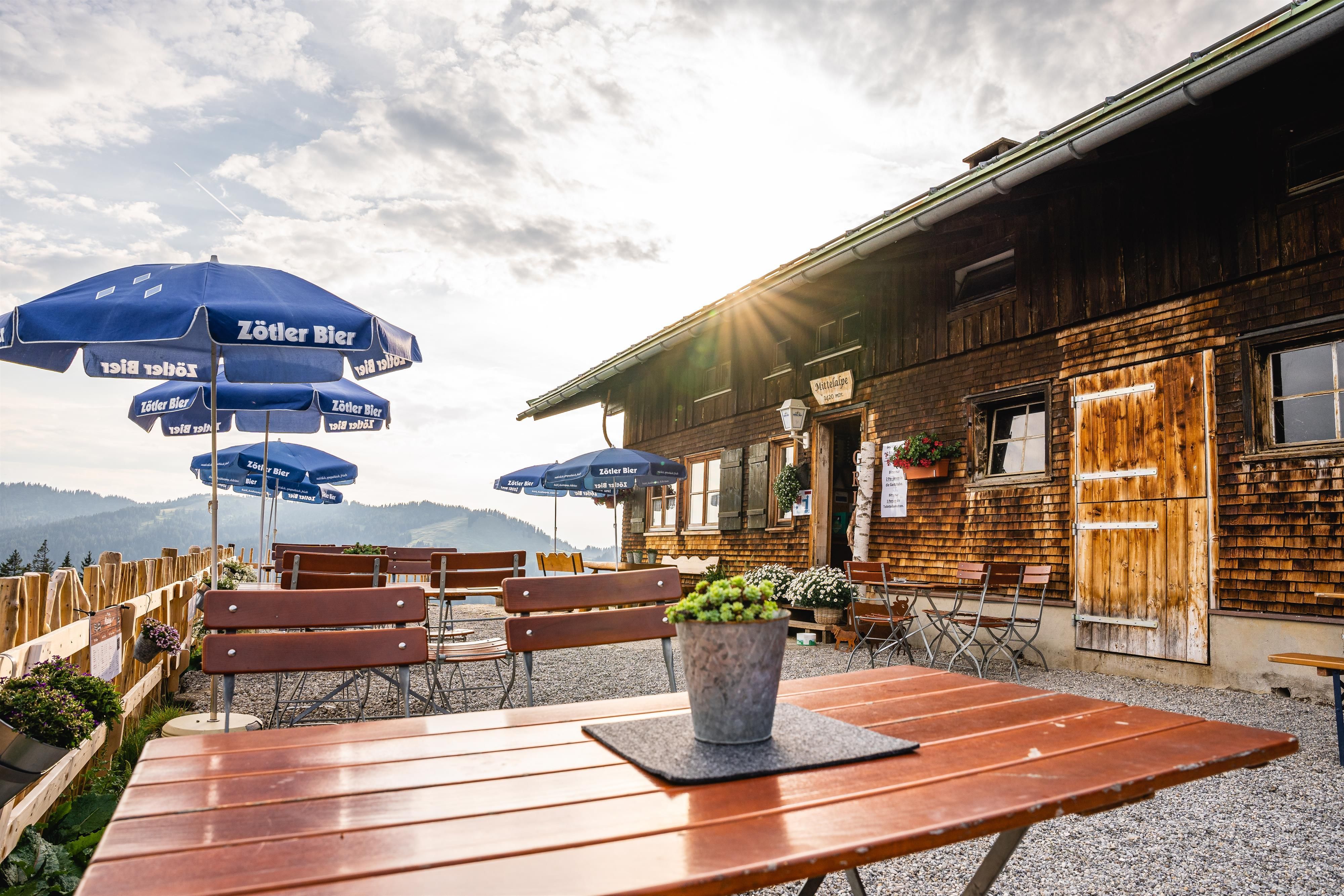 Mittelalpe im Sommer: Holzhaus mit Außenbereich, Tische, Bänke, Sonnenschirme "Zötler Bier", Blumenkästen, Bergpanorama, sonniger Himmel.