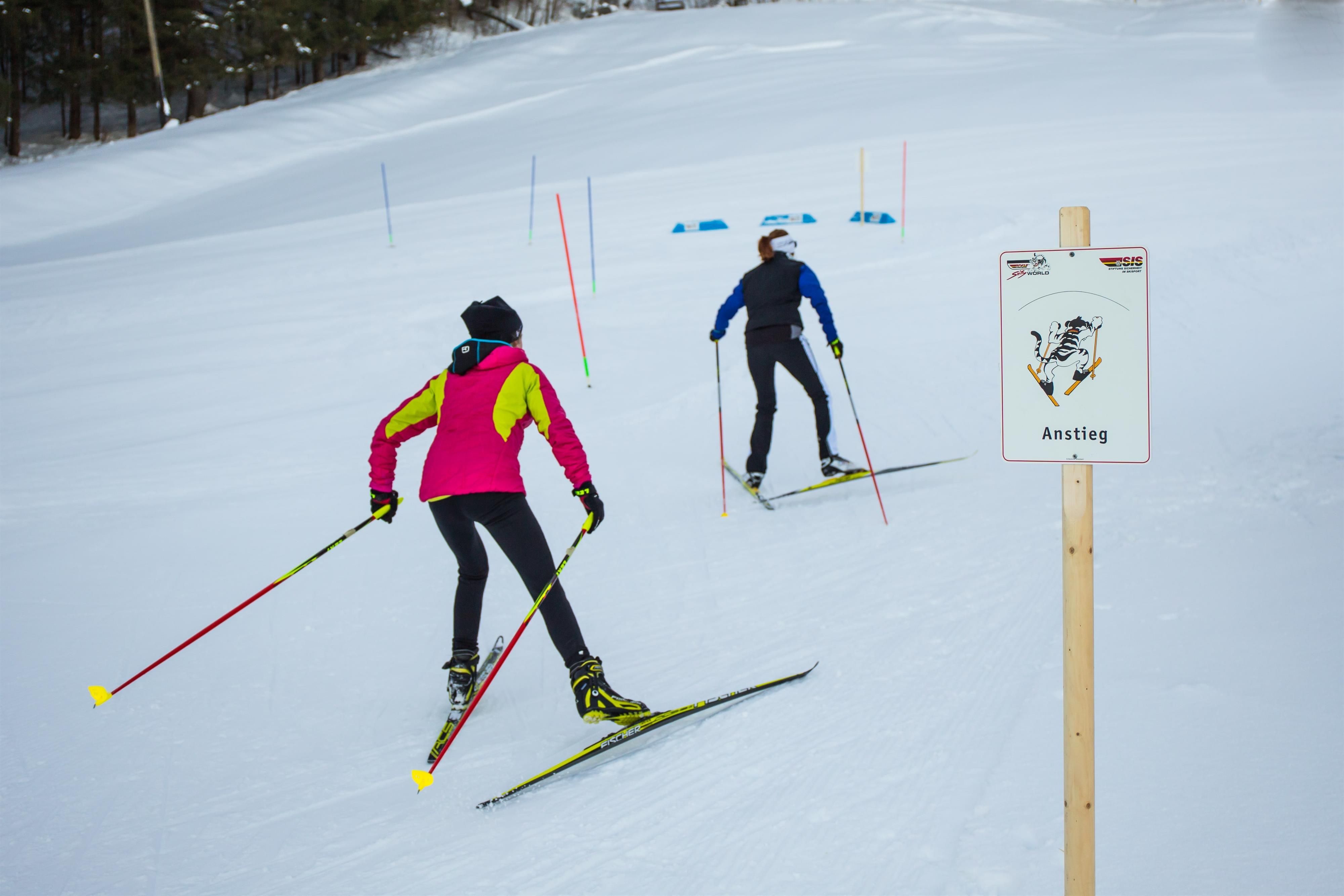 Zwei Langläufer auf Loipe, Anstiegsschild rechts, bunte Stangen markieren Strecke.