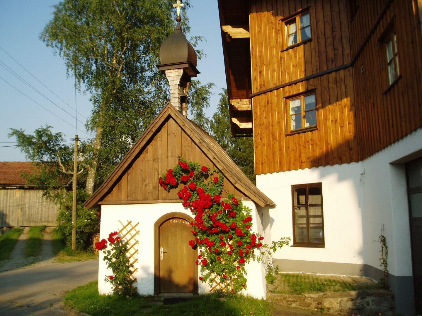 Kleine Kapelle mit Holzdach und -tür, umrankt von roten Rosen. Weißer Putzbau mit goldenem Kreuz auf dem Dach.