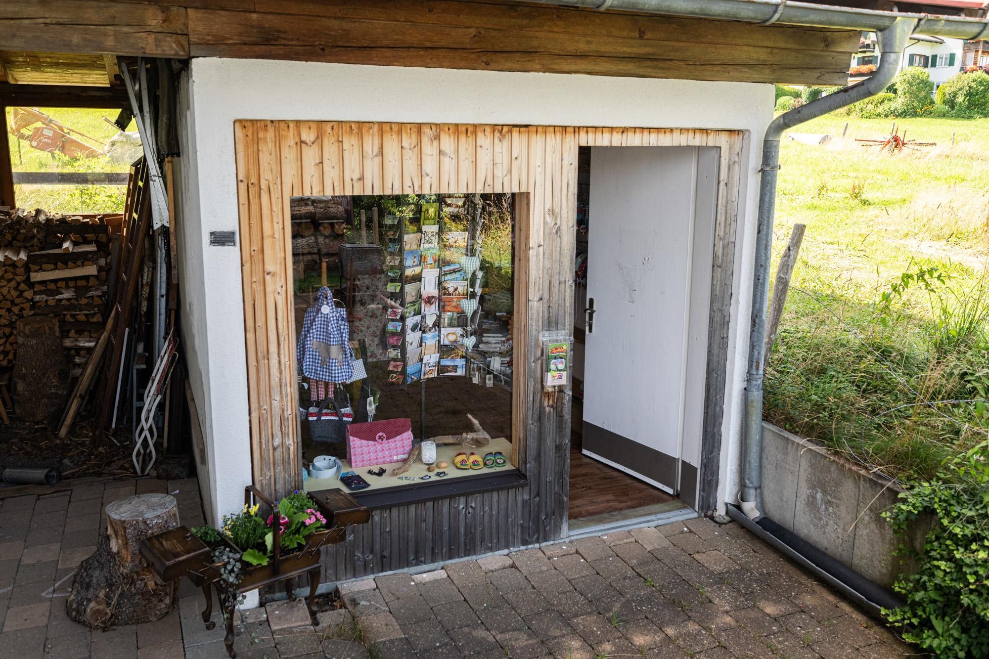 Kleiner Laden mit Schaufenster und offener weißer Tür an einem Gebäude mit Holzbalken und Steinpflaster.